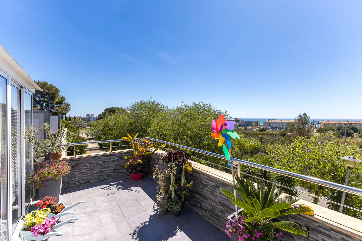 Balcony view with gray tile floor, stone wall, and metal railing. Colorful pinwheel, potted plants, and ocean view in the background.
