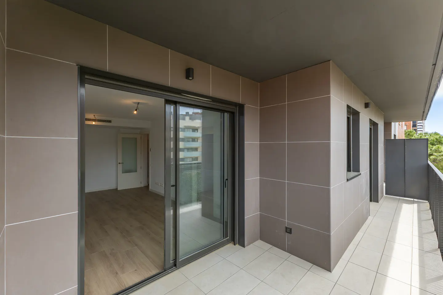 A modern apartment balcony with gray tiled walls, a sliding glass door, and a view of the interior and cityscape.