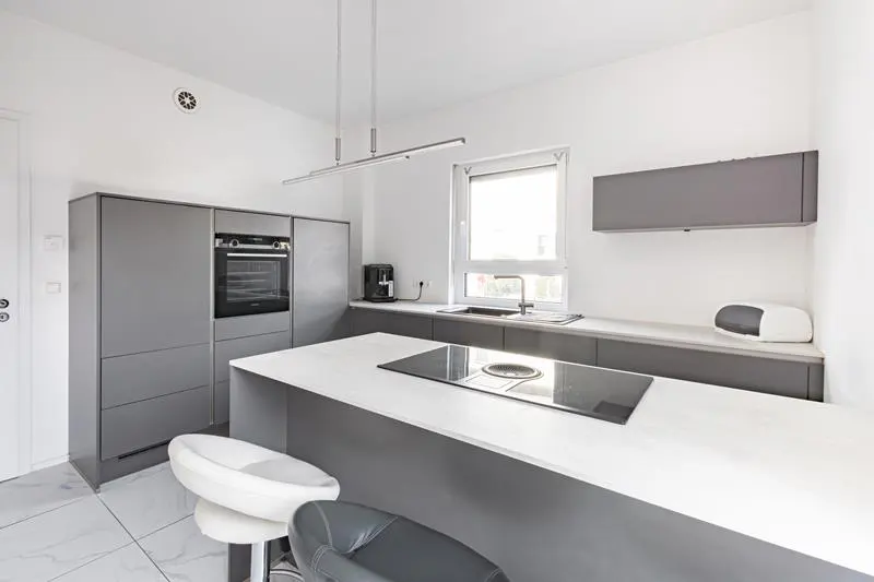 Modern kitchen with gray cabinets, white countertops, and island with cooktop. Two stools are in the foreground.