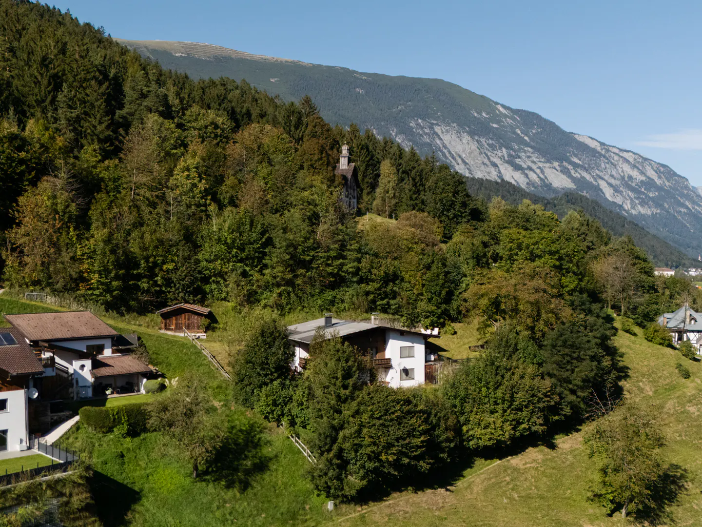 Scenic view of houses nestled on a green hillside with a backdrop of lush trees and a majestic mountain range under a clear blue sky.