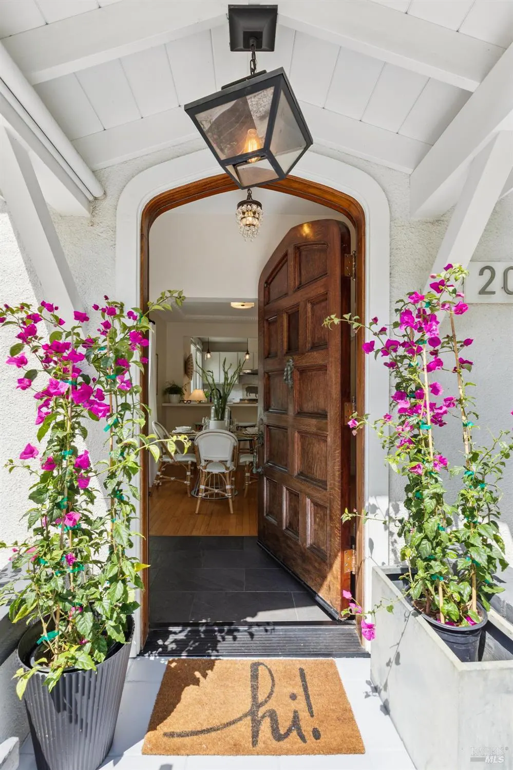 Open wooden front door with a "hi!" doormat, flanked by potted pink bougainvillea plants. Interior view of dining area with white chairs.