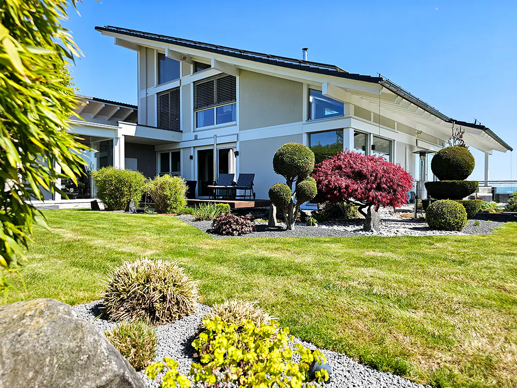 Modern two-story house with a manicured lawn, topiary trees, and a red maple tree under a clear blue sky.