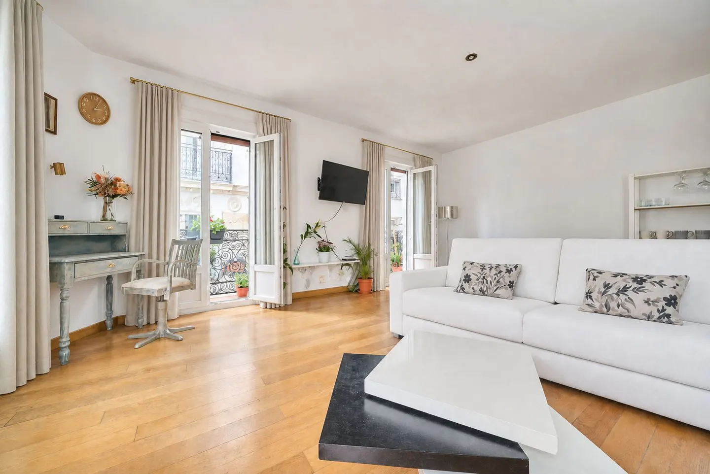 Bright living room with hardwood floors, white walls, and a white sofa. French doors open to a balcony. A vintage desk sits near the window.