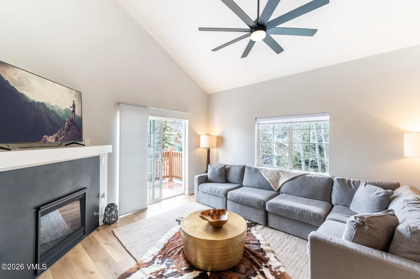 Living room with gray sectional sofa, gold coffee table, fireplace with TV, and a ceiling fan. Sliding glass doors lead to a deck.