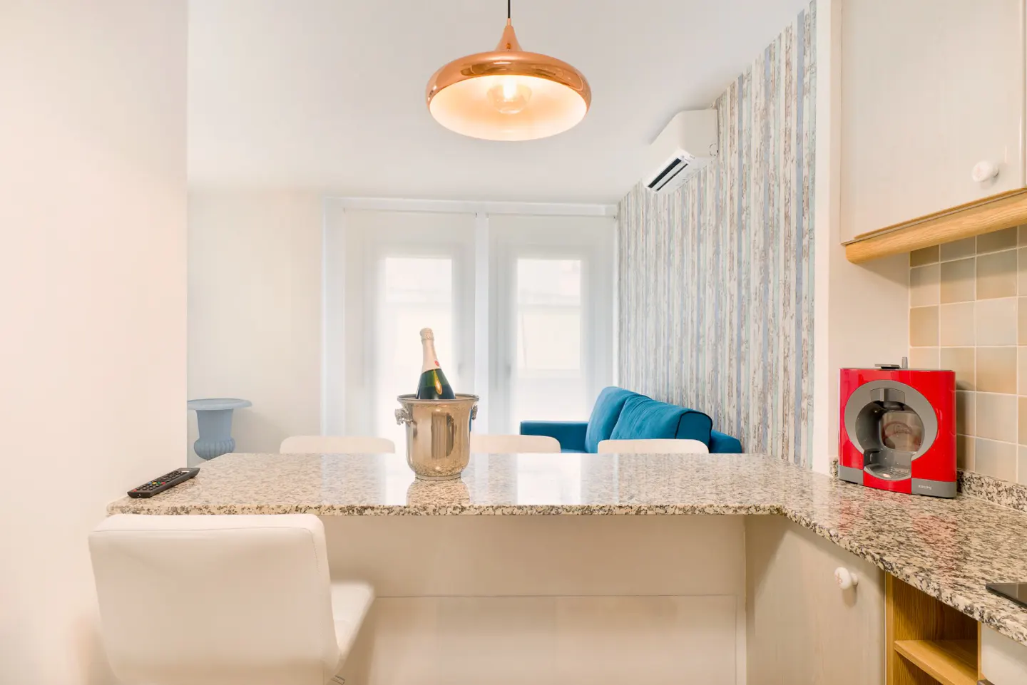Interior view of a modern apartment with a champagne bucket on a granite countertop, a blue sofa, and a copper pendant light.
