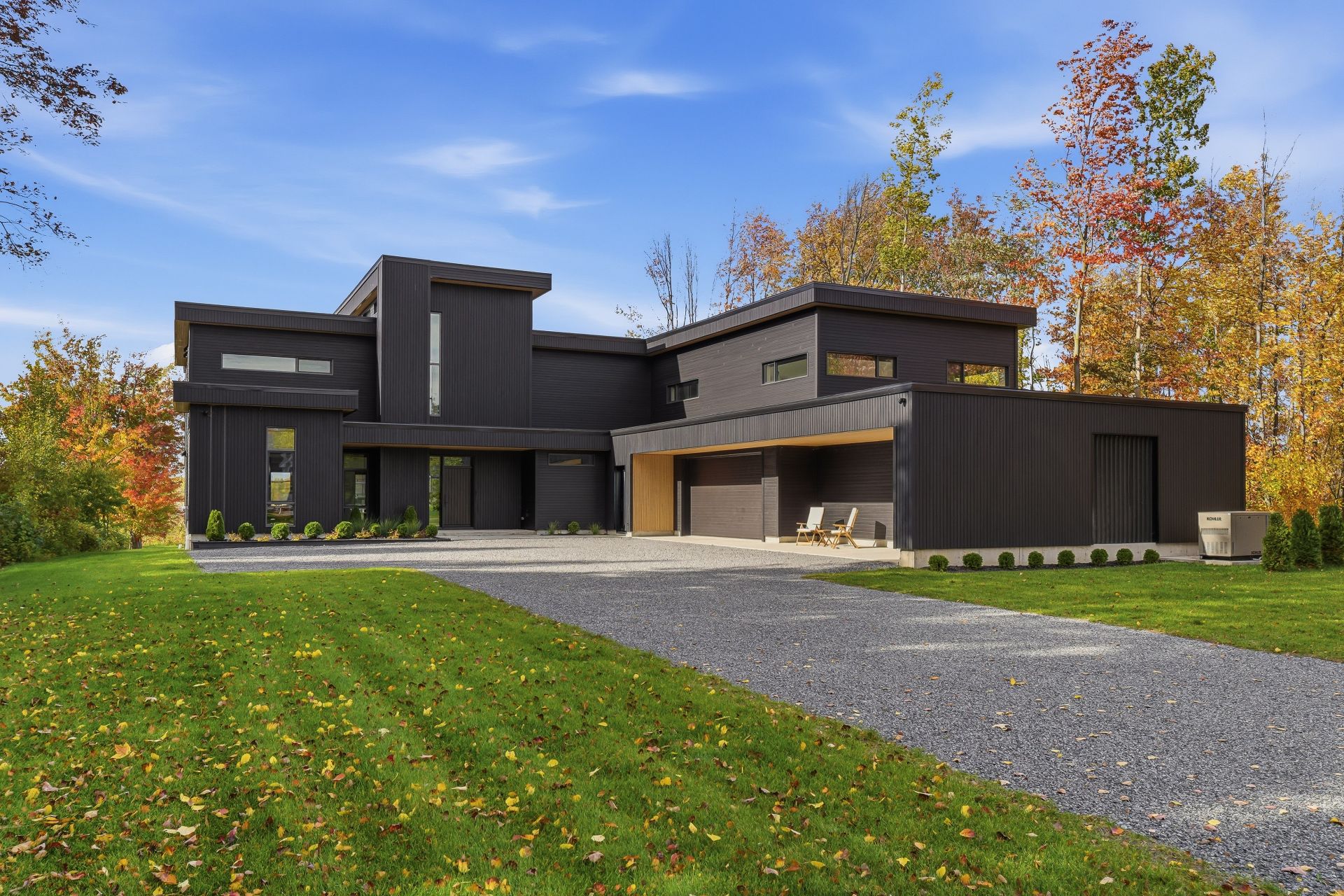 Modern black home with a gravel driveway, surrounded by autumn trees under a blue sky. [AI-generated text]