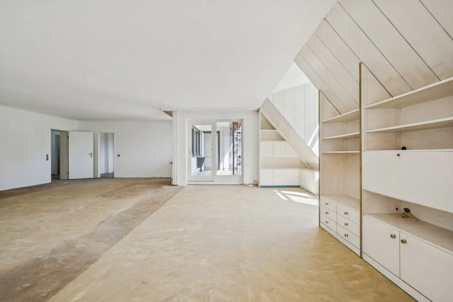 Bright, empty room with white walls, unfinished floor, and built-in white shelves. Doors lead to other rooms. Natural light fills the space.