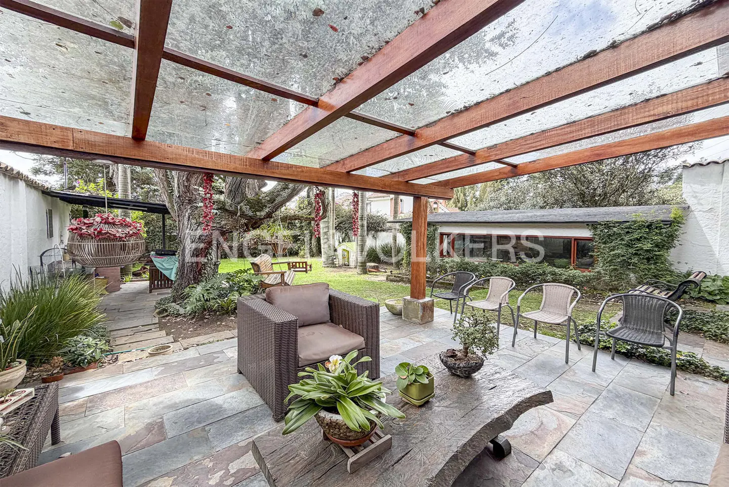 Outdoor patio with brown wicker furniture, stone flooring, and a clear pergola roof. Lush greenery and a building are in the background.