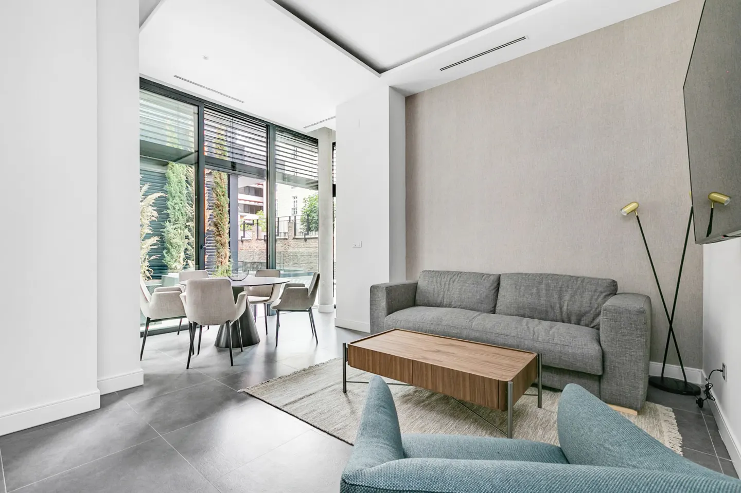 Bright living room with a gray sofa, wood coffee table, and blue chair. A dining table sits by large windows overlooking a canal.