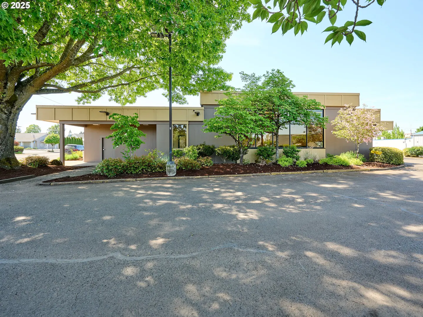 Exterior view of a one-story commercial building with a tan facade, large windows, and a drive-through. Trees and landscaping surround the building.