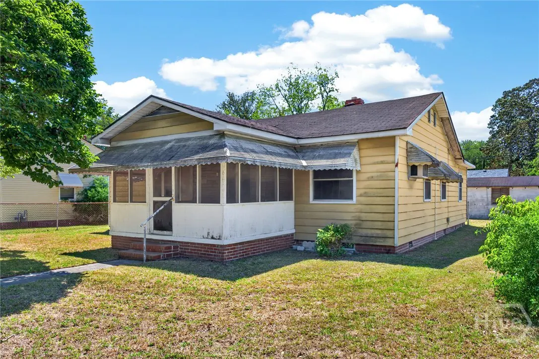 Exterior view of a one-story yellow house with a screened-in porch and a brown shingled roof.