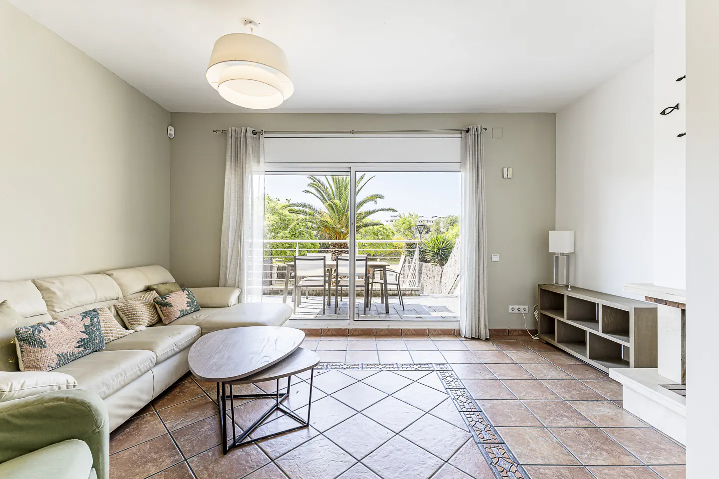 Living room with a white sofa, pillows, and a two-tiered coffee table. Sliding glass doors lead to a balcony with a table and chairs.
