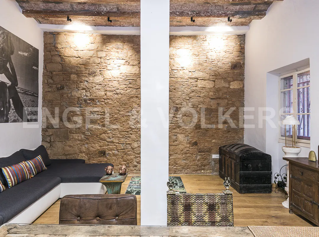 Living room with exposed stone walls, wooden floors, and a black sectional sofa. A large trunk sits near a window.