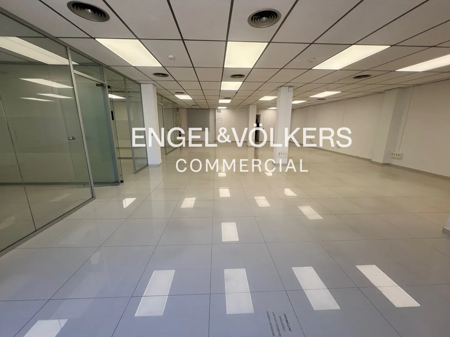 Empty commercial space with glass-walled offices, white walls, and gray tiled floors. The ceiling has recessed lighting and vents. "Engel & Volkers Commercial" is overlaid.