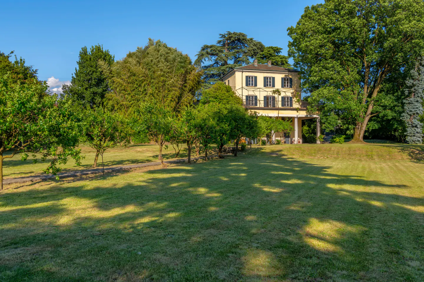 Two-story yellow house with black shutters, surrounded by green trees and a large lawn under a blue sky.