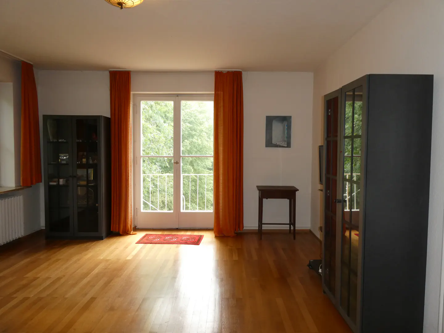 Living room with hardwood floors, orange curtains, glass-front cabinets, a small wooden table, and a balcony door.