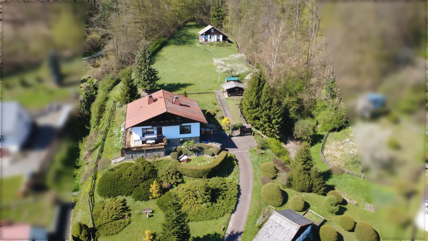 Aerial view of a white house with a red roof, surrounded by green trees and grass. A driveway leads to the house.
