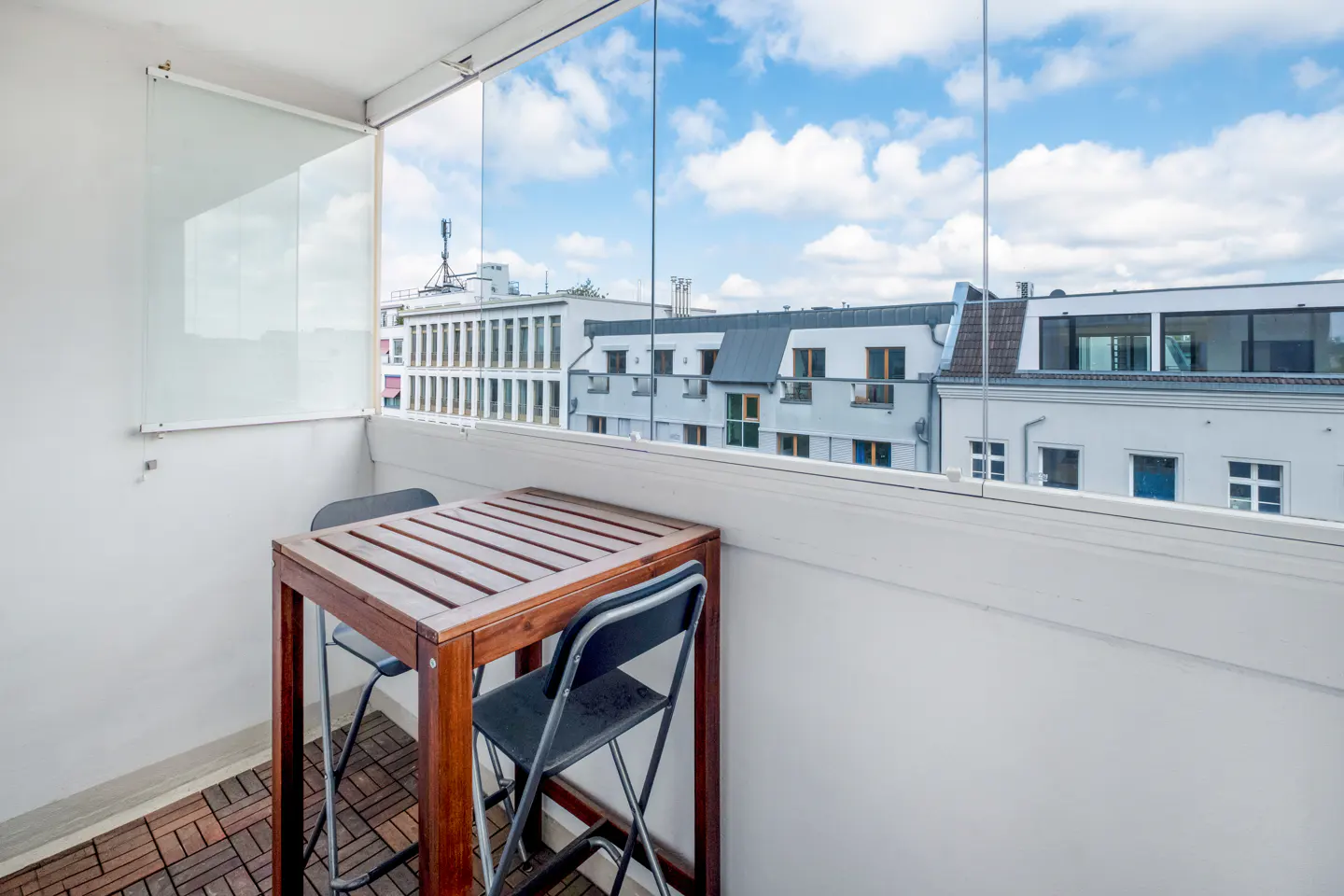 Balcony with a wooden table and two chairs, glass walls, and a view of white buildings under a blue sky.