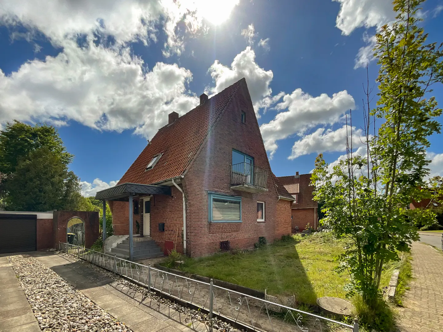 A two-story brick house with a red tile roof under a sunny, cloudy sky. A small balcony is visible on the second floor.