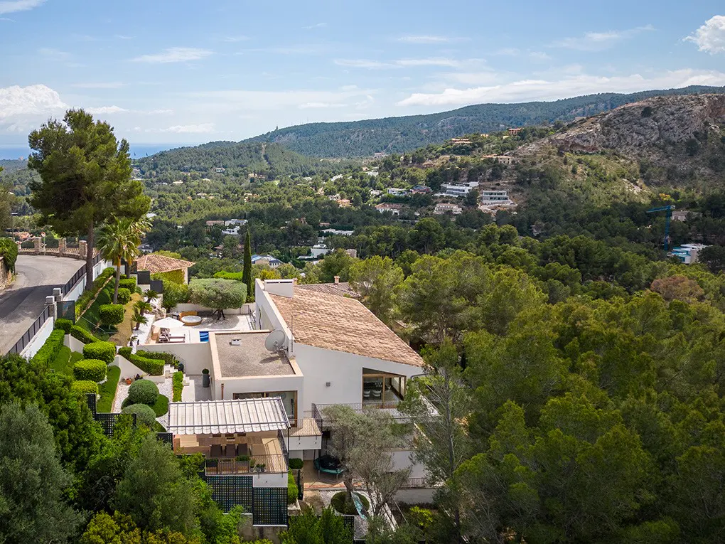 Aerial view of a modern white house with a brown tile roof, surrounded by lush green trees and hills under a blue sky.