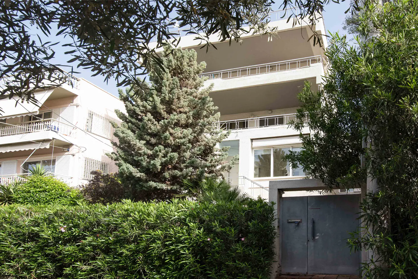 Exterior of a white, multi-story building with balconies, surrounded by green trees and bushes, and a gray gate.