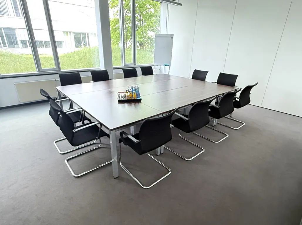 Bright conference room with a large wood table, black chairs, and a tray of drinks. A whiteboard stands near a window with a green view.