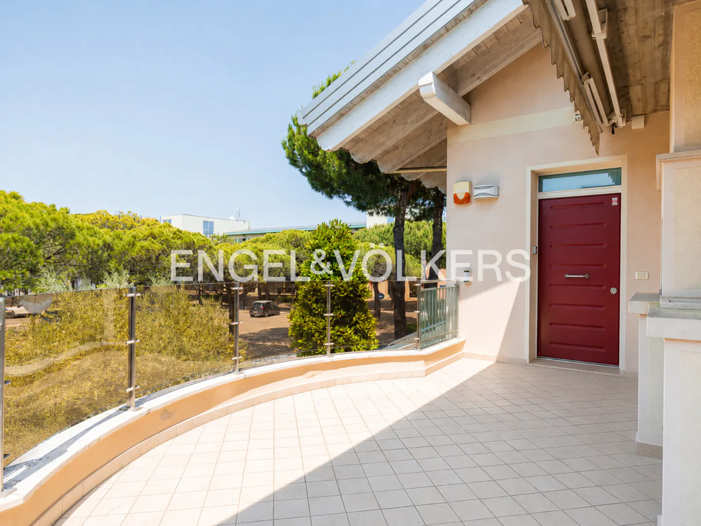 Exterior view of a house with a red front door and a tiled patio with a glass railing. Trees are visible in the background.