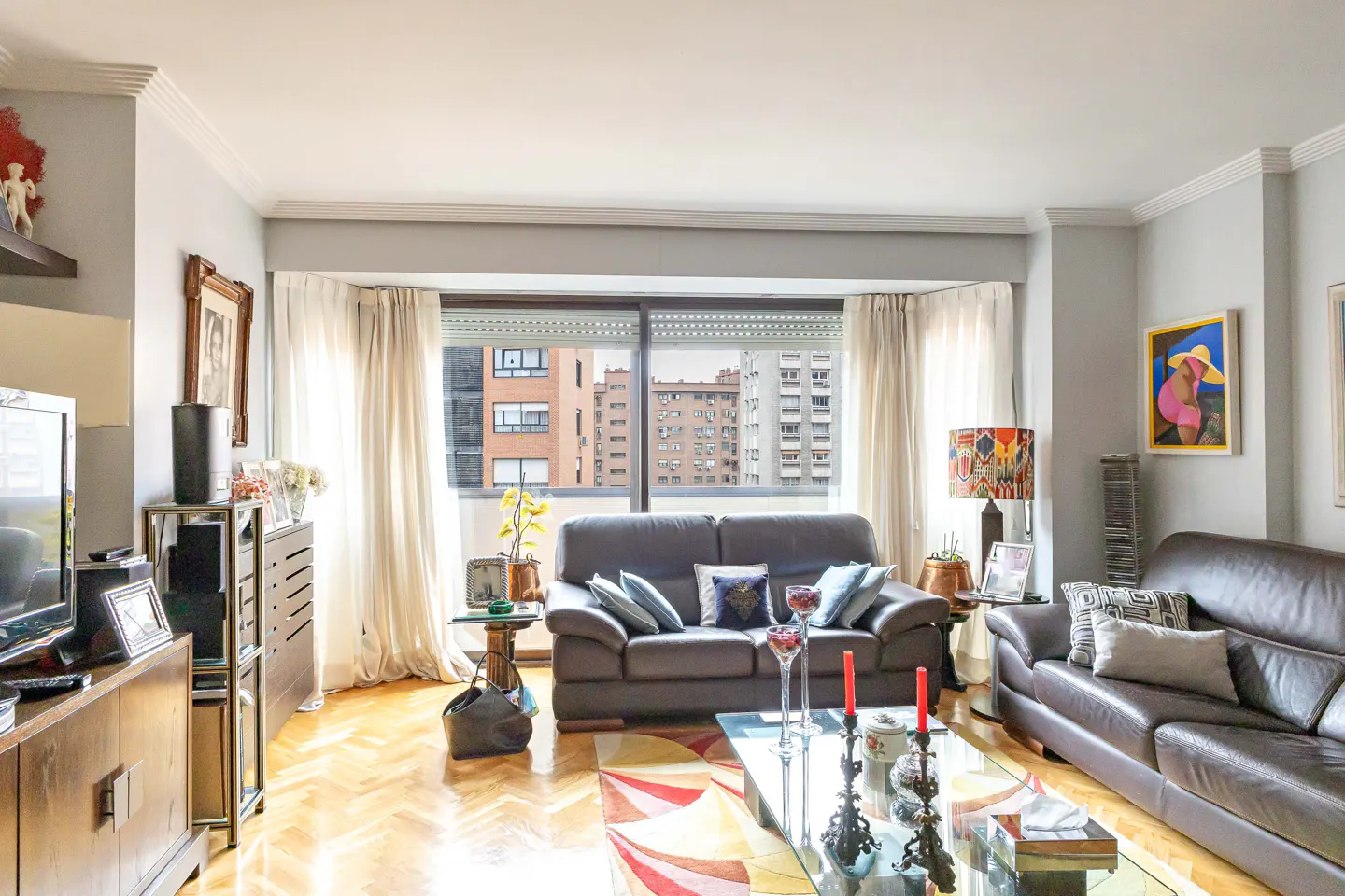Bright living room with two dark leather sofas, a glass coffee table, hardwood floors, and a large window with city views.