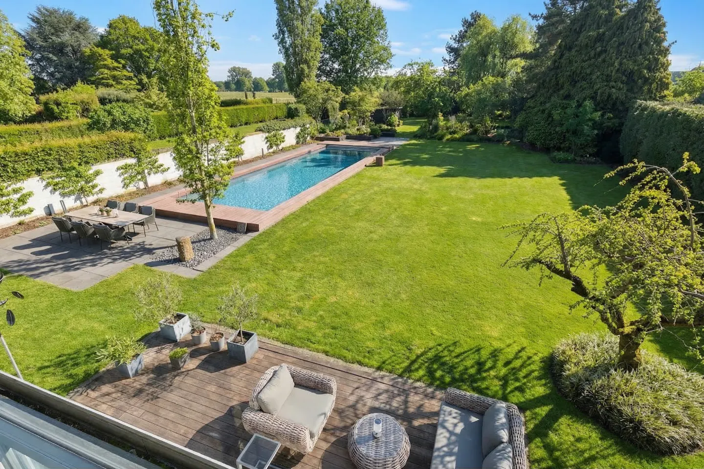 Aerial view of a backyard with a pool, patio dining set, and a wooden deck with wicker furniture. Green lawn and trees surround the area.