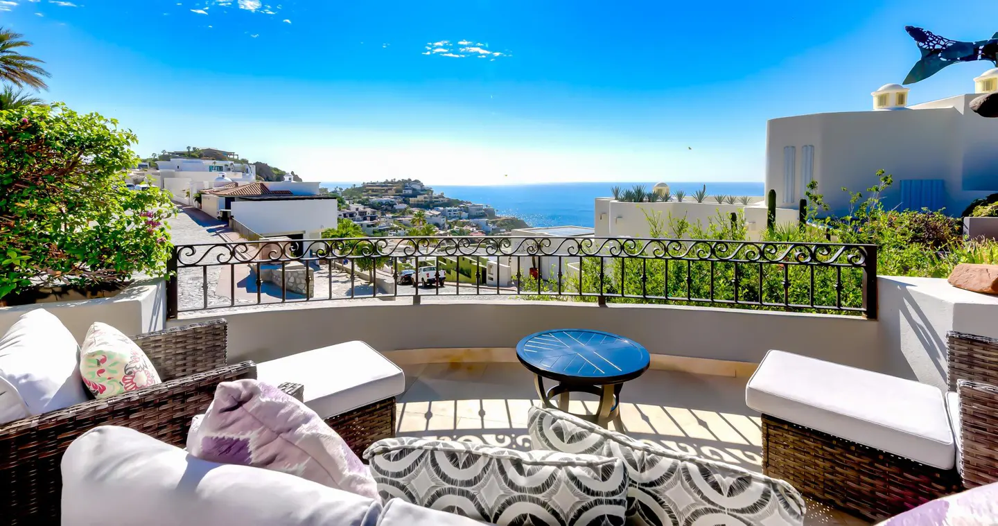Balcony view with wicker furniture, pillows, and a blue table overlooking a coastal town and ocean under a bright blue sky.