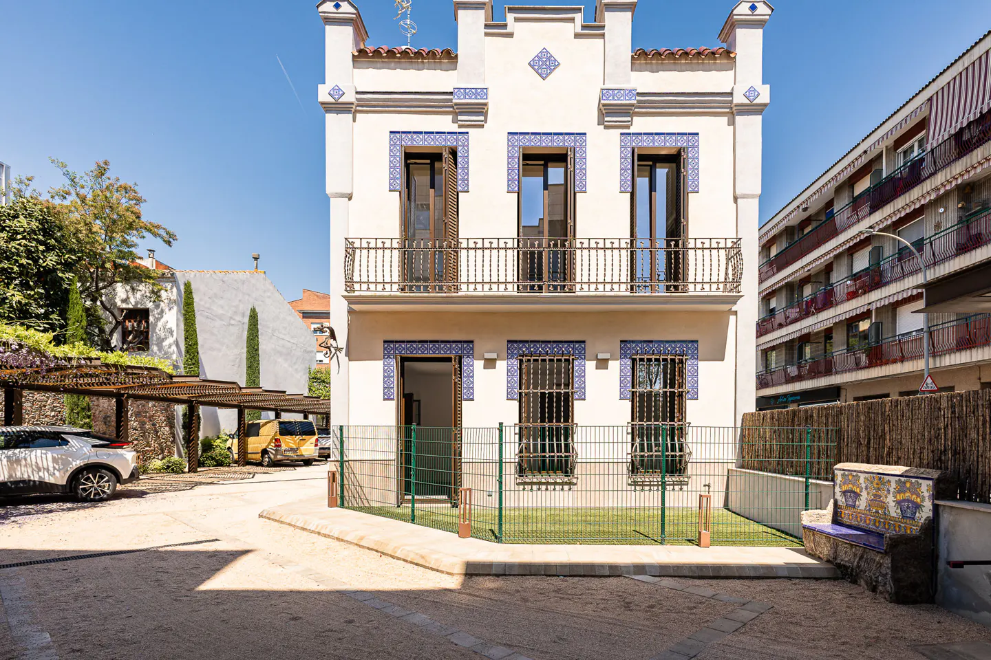 Two-story white building with blue tile accents, brown shutters, and a small balcony. A green fence surrounds a small lawn. Cars are parked under a pergola.