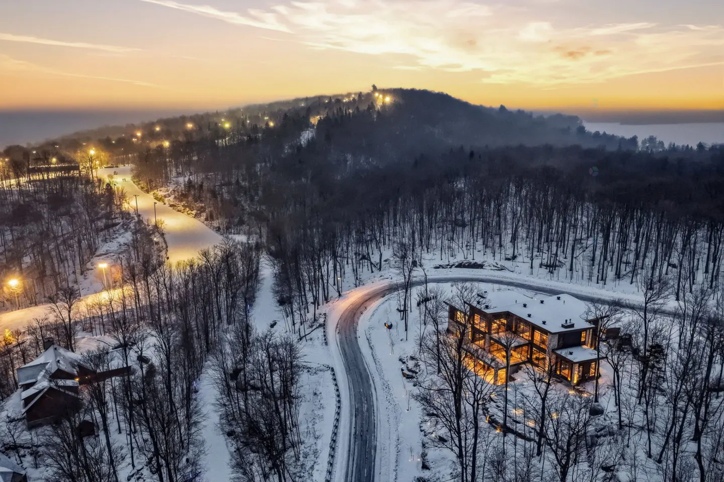 Aerial view of a modern house with lights on, surrounded by snow-covered trees and a winding road at dusk.
