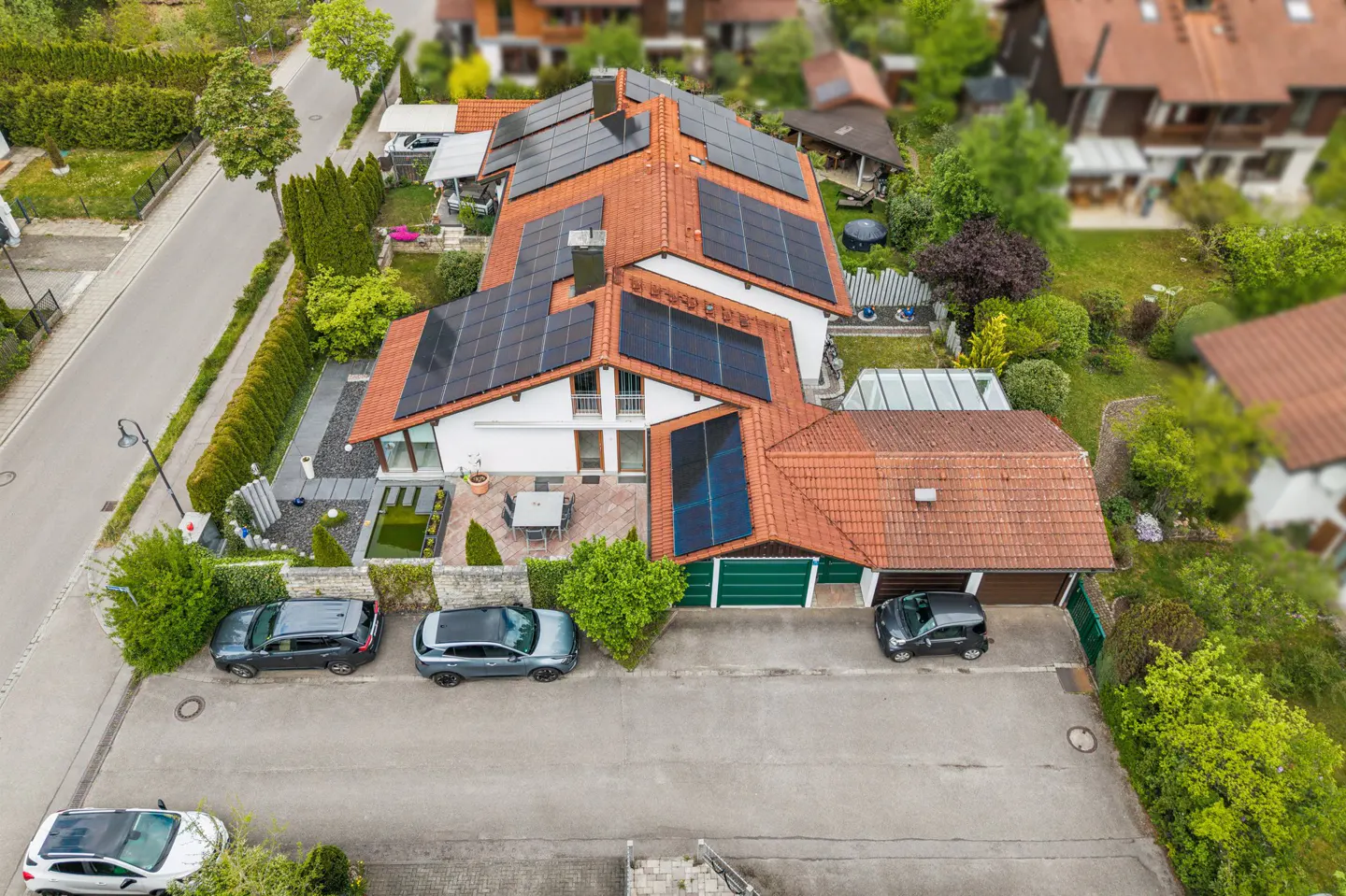 Aerial view of a white house with a red tile roof covered in solar panels, a green lawn, and three cars parked in front.