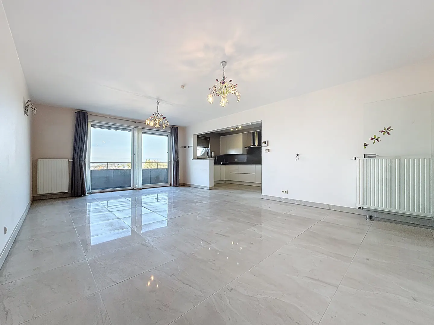 Bright, empty apartment with white walls, marble floors, and a modern kitchen. Chandeliers hang from the ceiling. Balcony with city view.