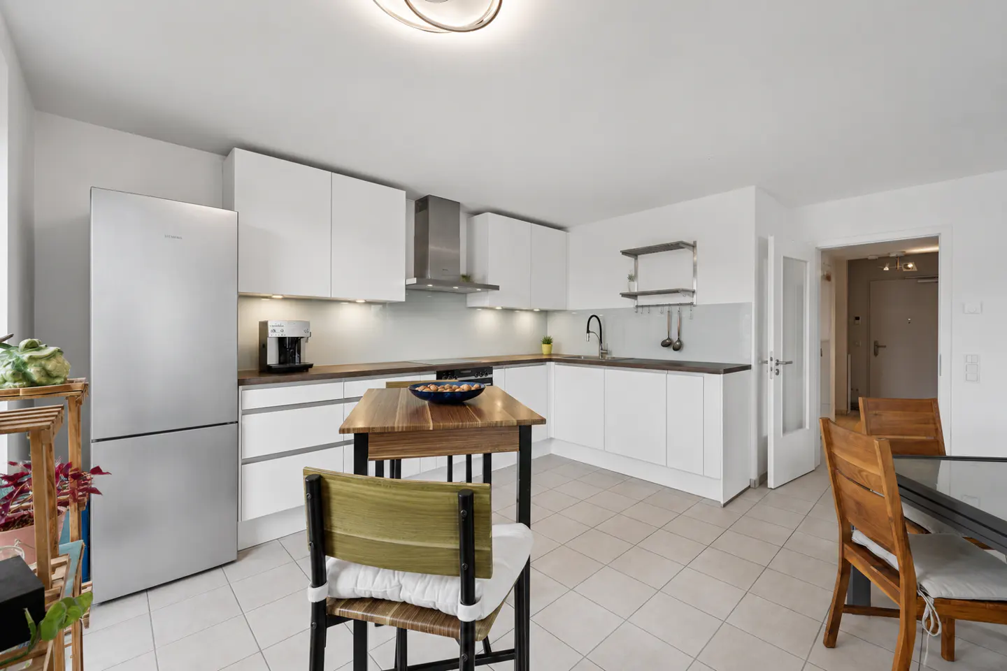 Bright, modern kitchen with white cabinets, stainless steel appliances, and a wooden table with chairs. Tiled floor and open doorway visible.