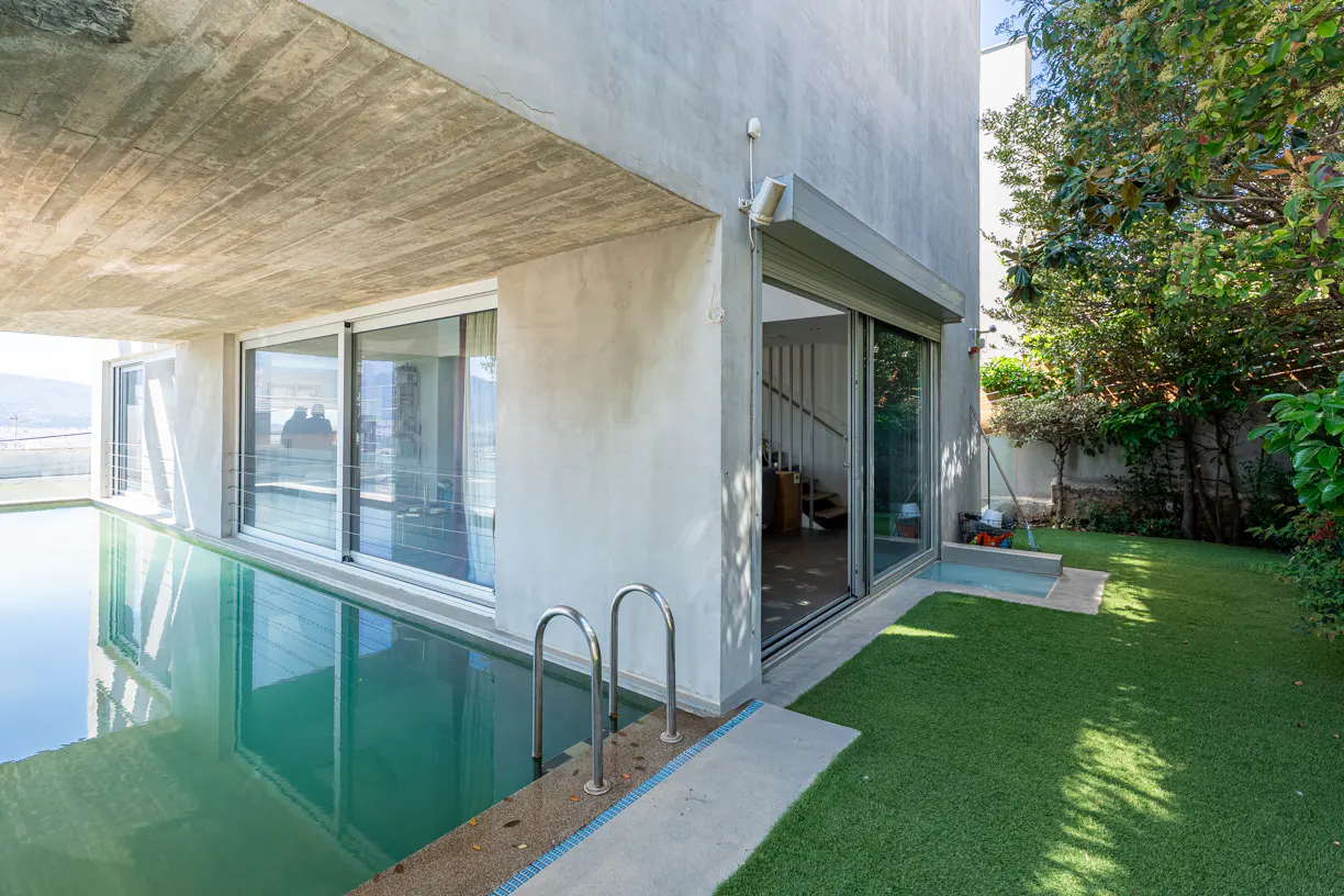 Exterior view of a modern home with a pool, sliding glass doors, and a green lawn. The building is concrete and has a flat roof.