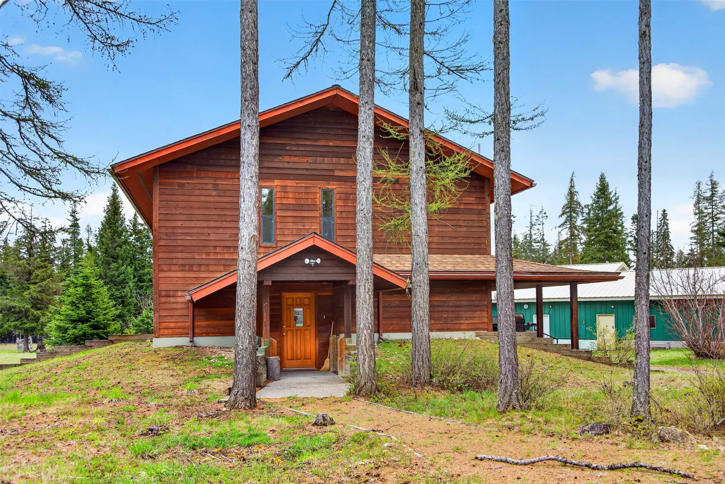 Two-story brown wood house with orange trim, seen through tall trees on a grassy lawn. Blue sky in the background.