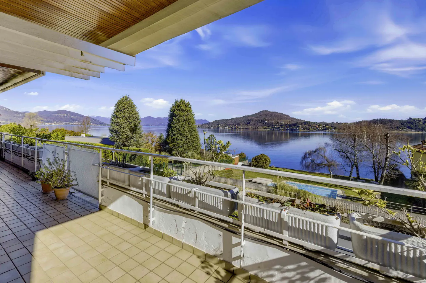 Balcony view of a lake with mountains in the background. White railings and planters line the balcony. Blue sky with clouds above.