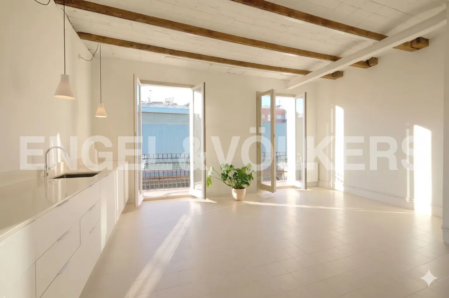 Bright, airy room with white walls, wood beams, and open doors to balconies. A white kitchen counter is on the left. A potted plant sits near the balcony doors.