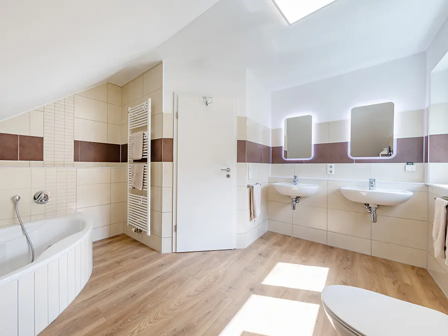 Bright bathroom with wood floors, white walls with tan and brown tile accents, two sinks with lighted mirrors, and a white tub.