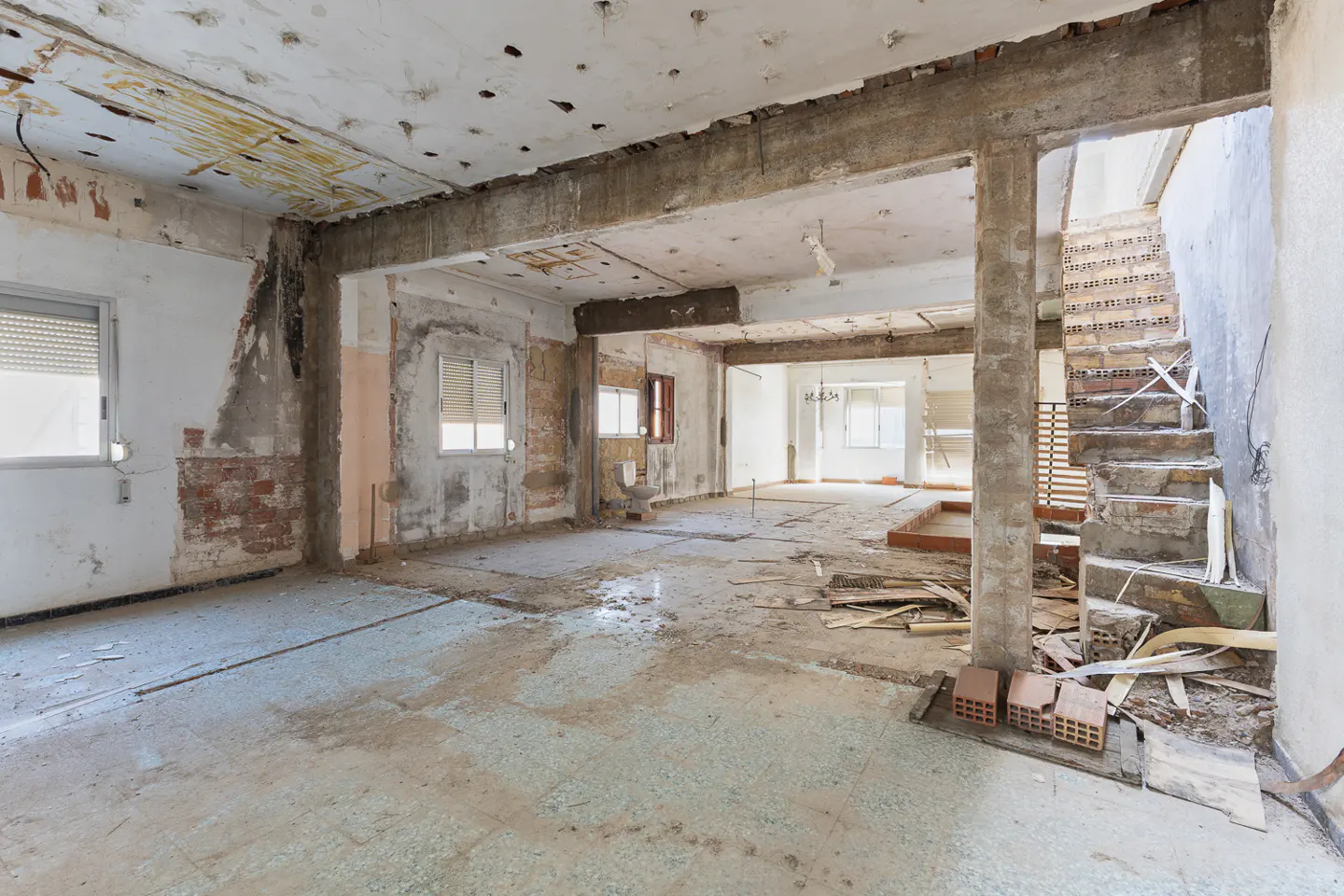 Interior of a dilapidated house with exposed brick, concrete beams, and debris on the floor. Stairs lead to an upper level.