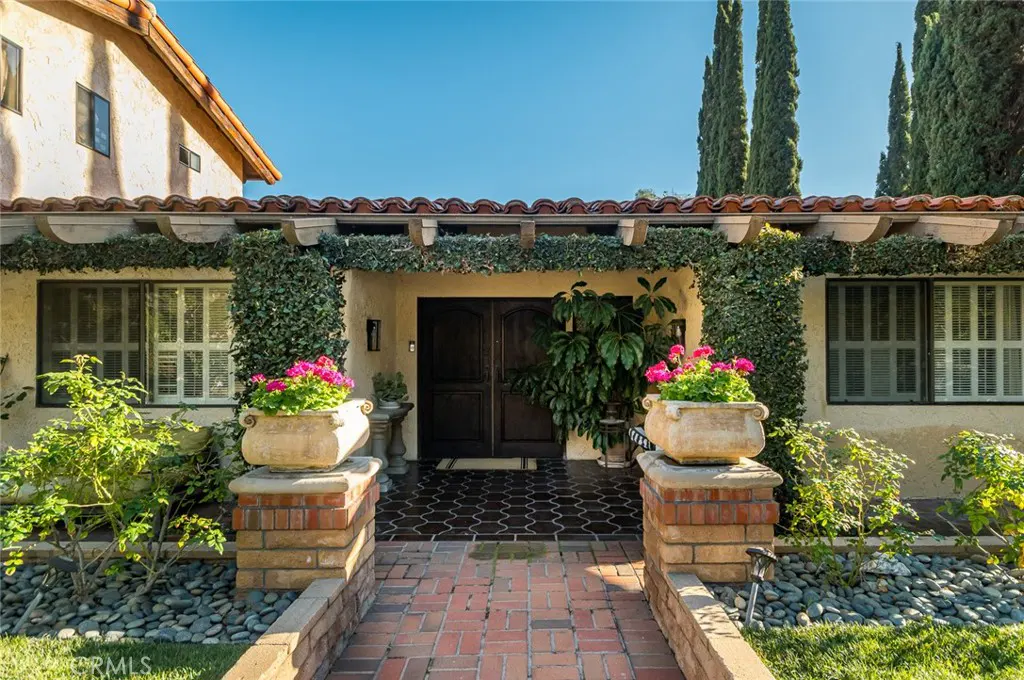 Front entrance of a Spanish-style home with a brick walkway, potted pink flowers, and dark wood double doors.