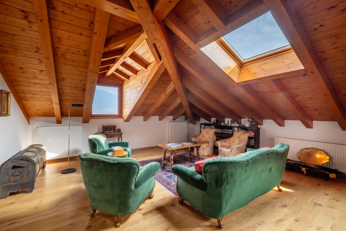 Attic living room with wood floors, exposed beams, and a skylight. Green velvet sofas and chairs surround a wooden coffee table on a patterned rug.