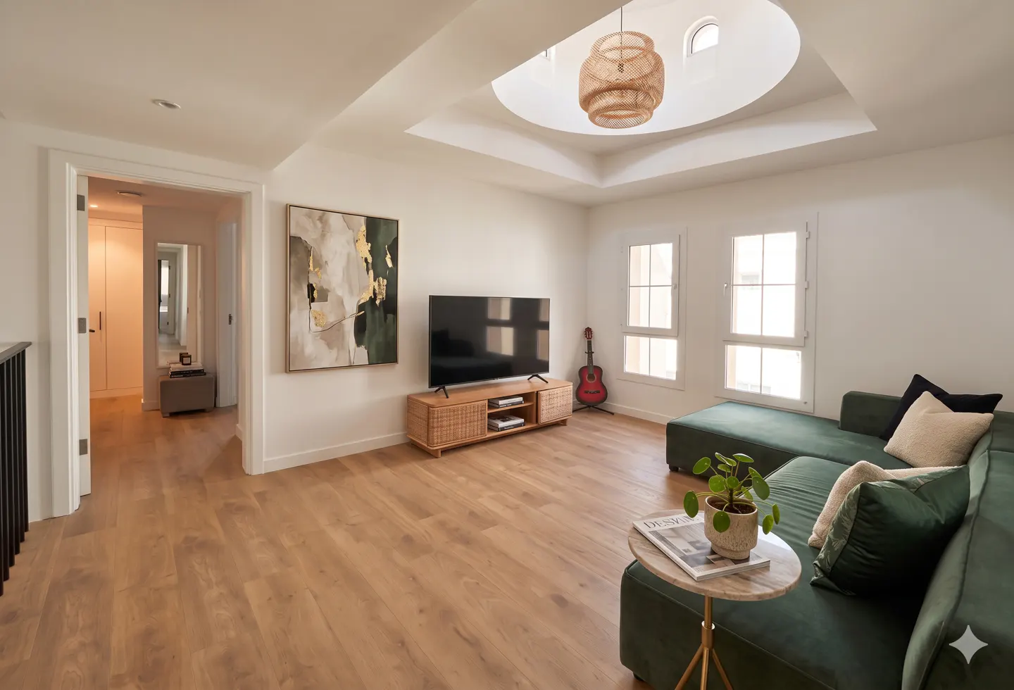 Bright living room with wood floors, white walls, and a green sectional sofa. A TV sits on a woven console, with a guitar nearby. A round skylight illuminates the space.