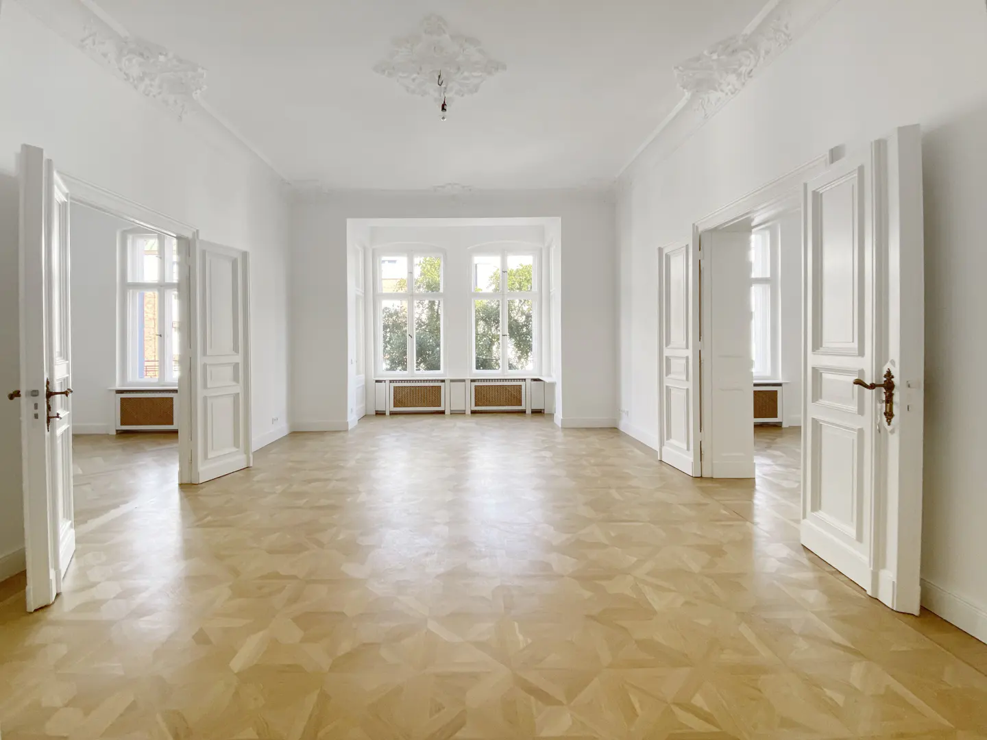Bright, empty room with white walls, light wood floors, and open white doors. Windows let in natural light.