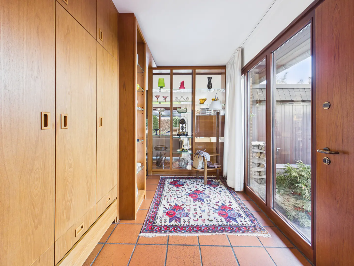 Hallway with wood cabinets, glass display case, and a patterned rug on terracotta tile floor. A glass door leads to an outdoor area.
