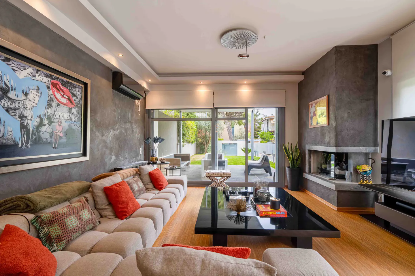 A modern living room with a large beige sofa, orange pillows, and a black coffee table. A large painting hangs on the gray wall. Sliding glass doors lead to a patio.