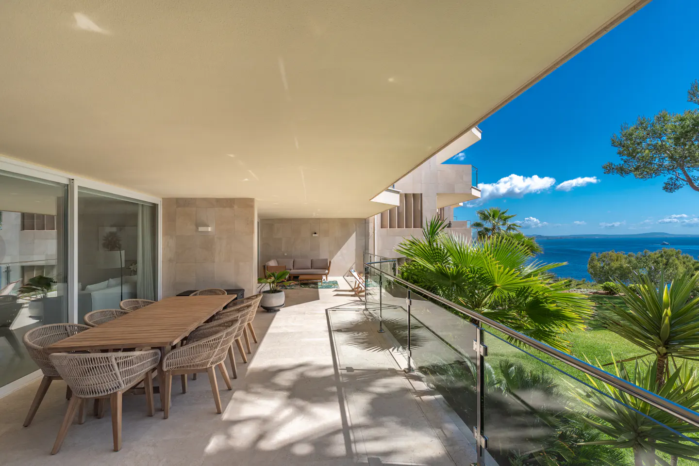 A sunlit patio with a long wooden table and woven chairs overlooks a blue ocean and lush green foliage.