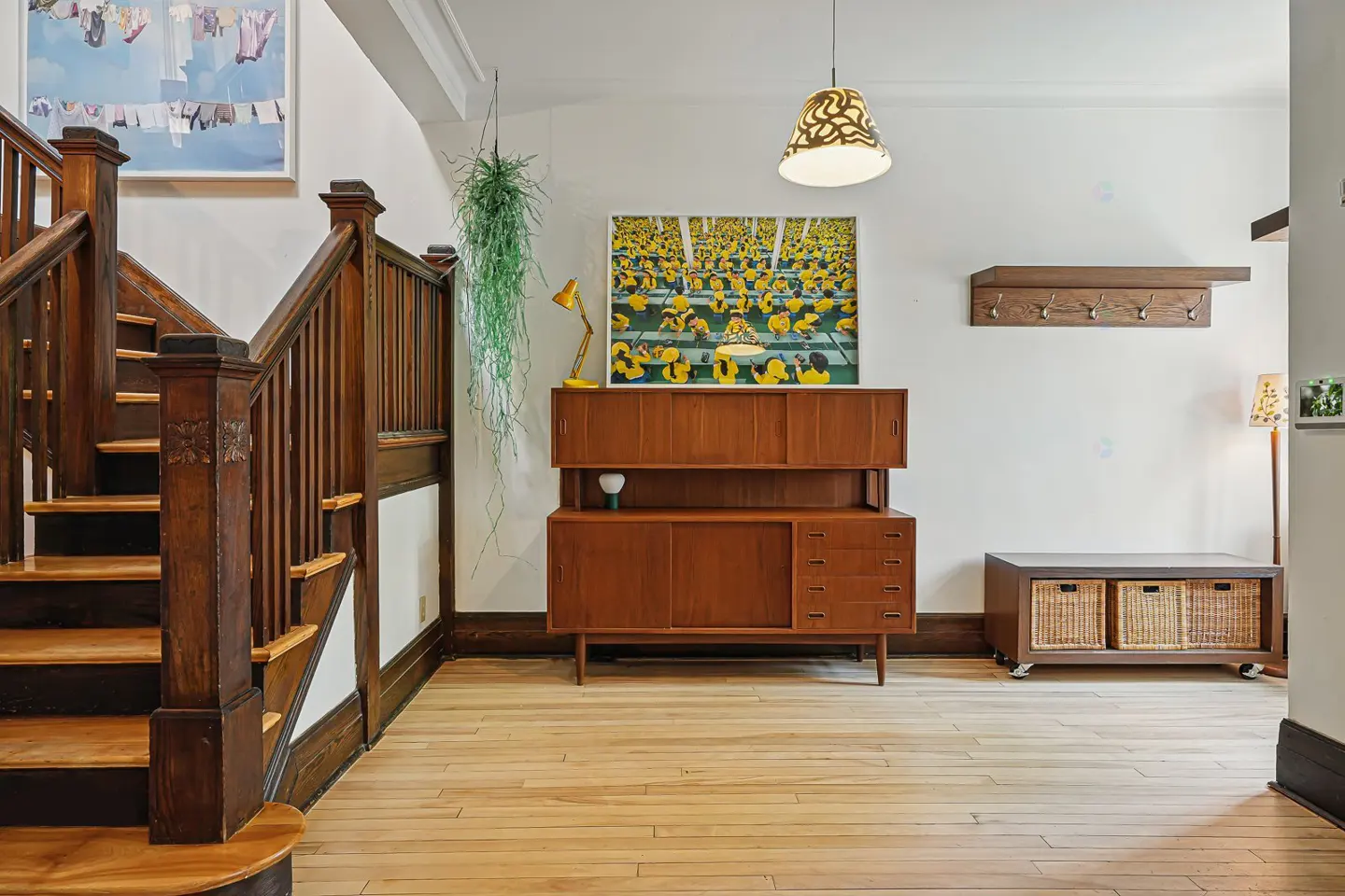 Foyer with wooden staircase, light wood floors, and mid-century modern furniture. Artwork and hanging plant add color.