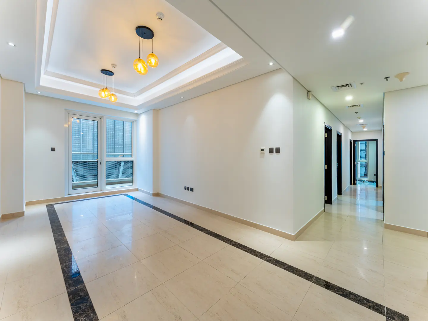 Bright, empty room with beige tile floor, white walls, and modern lighting fixtures. A hallway with dark doors is visible.